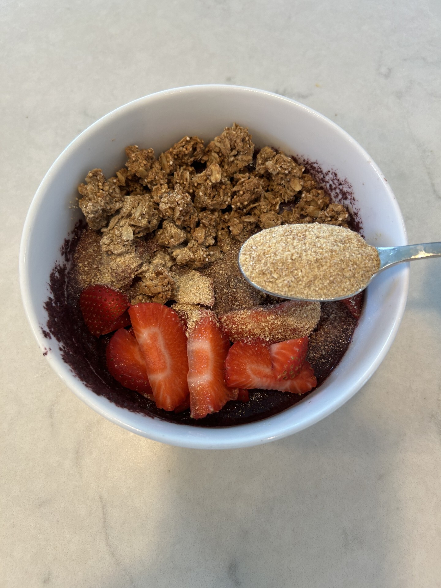 Blueberry smoothie bowl topped with sliced strawberries, granola and wheat germ, with a spoonful of wheat germ above the bowl.